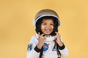 Delighted little African American girl in white space suit touching helmet while looking away on orange background