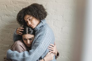 Sad African American woman with closed eyes hugging and consoling depressed female friend near white wall in light room at home